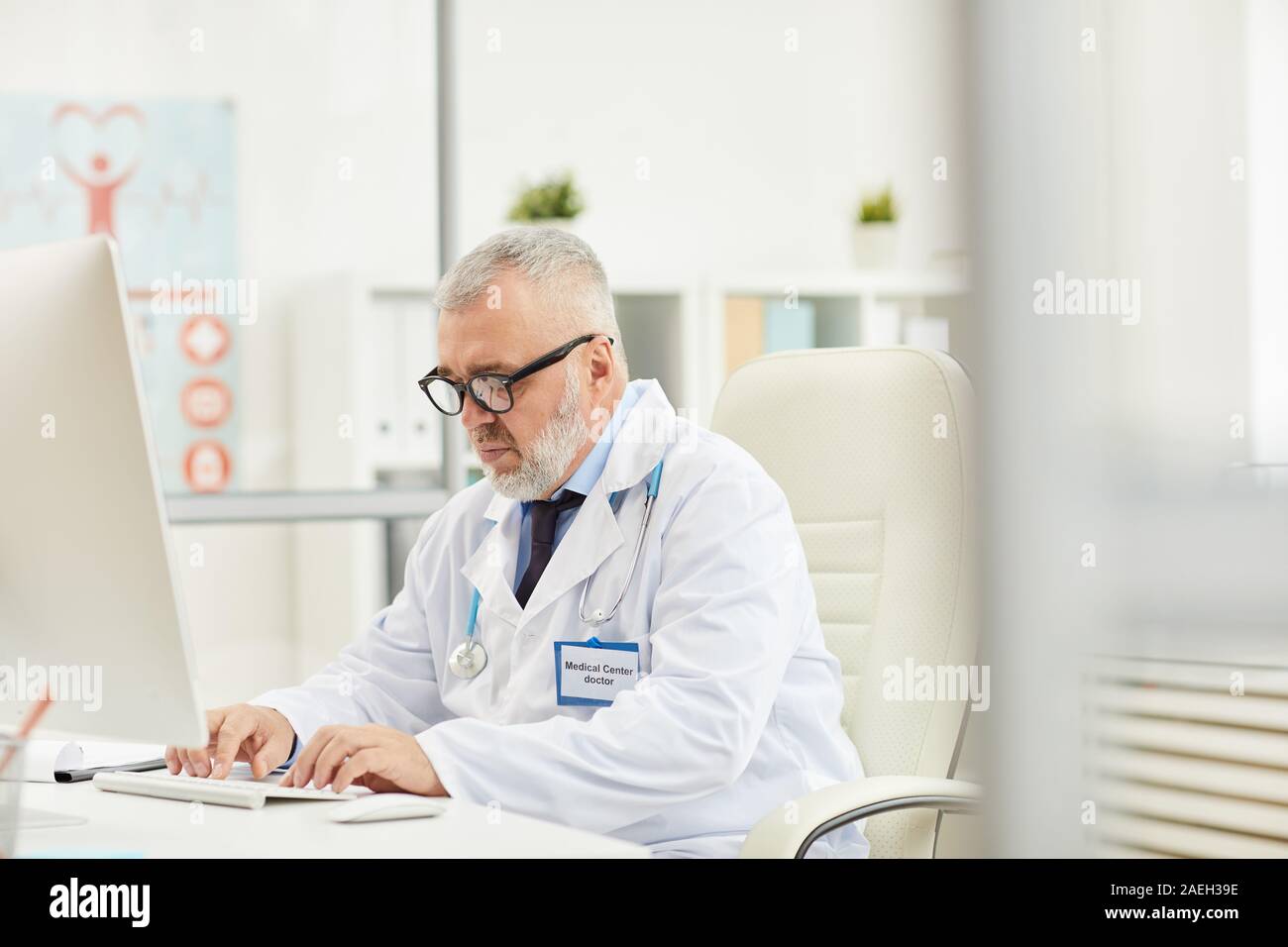 Serious mature medical professional sitting at the table and working on computer at his office Stock Photo