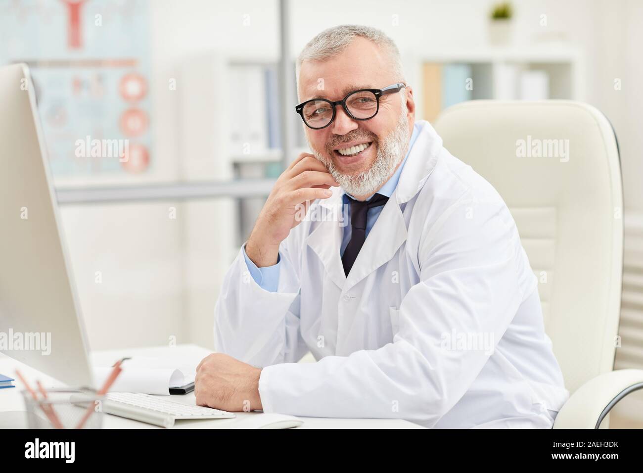 Portrait of successful mature doctor in eyeglasses sitting at his workplace and smiling at camera at office Stock Photo