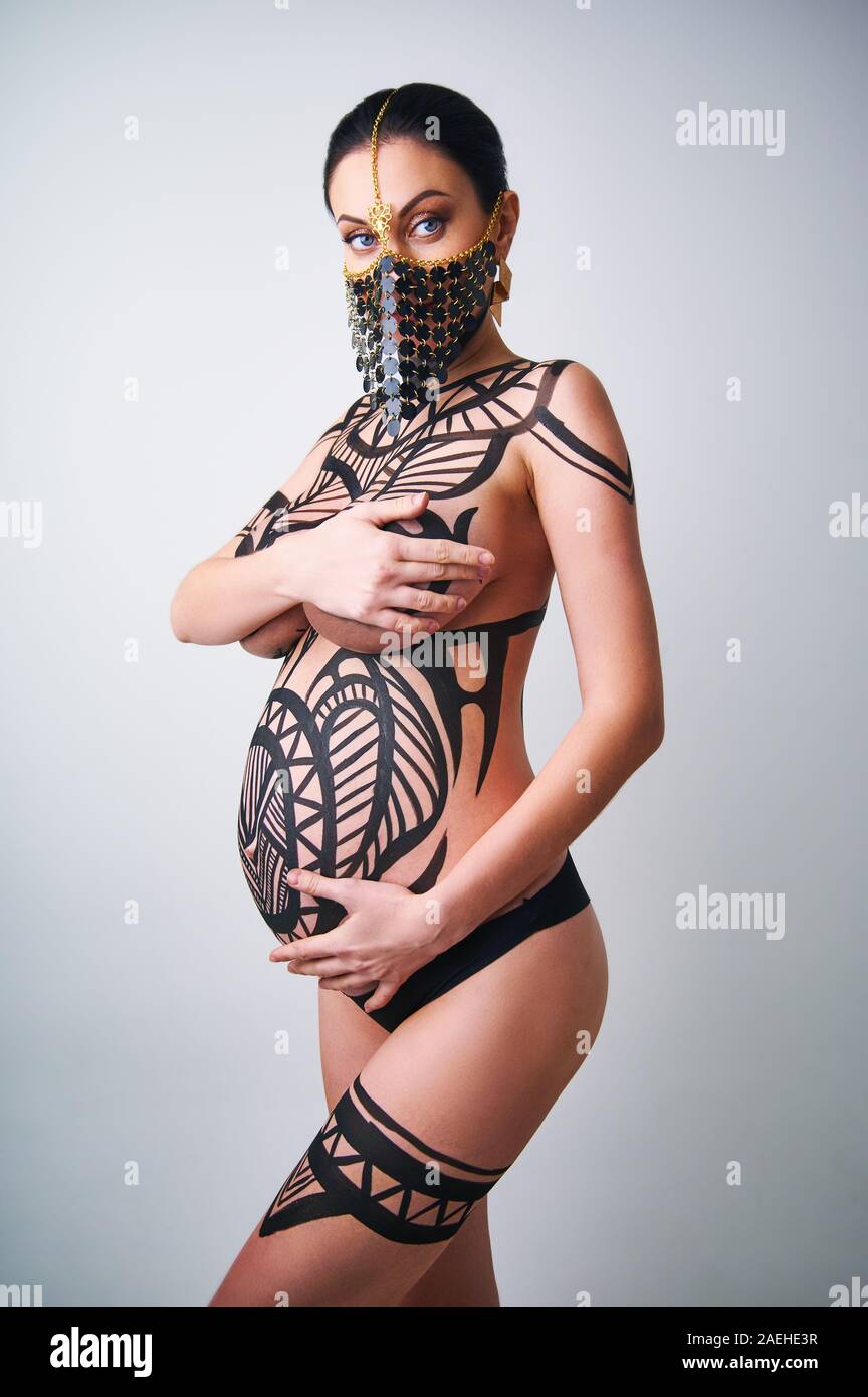 Naked pregnant woman with painted body art. Woman's face half hidden by a ornament. Studio photo Stock Photo