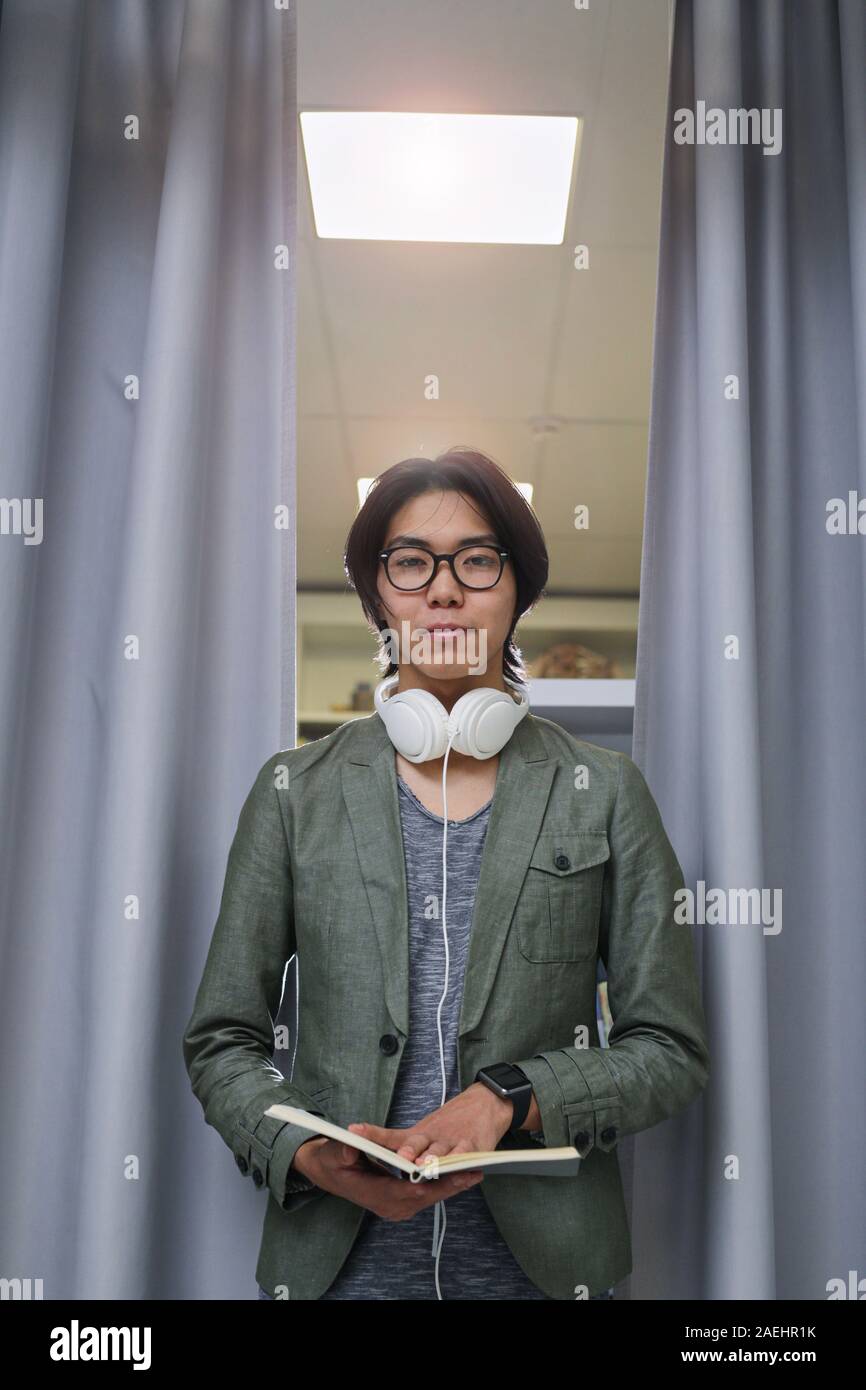 Portrait of Asian young man in headphones standing and looking at camera while reading a book Stock Photo