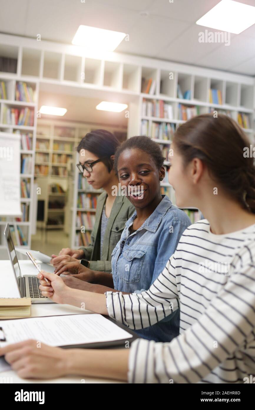Two young women talking to each other and smiling while using laptop at the table with young man working in the background Stock Photo