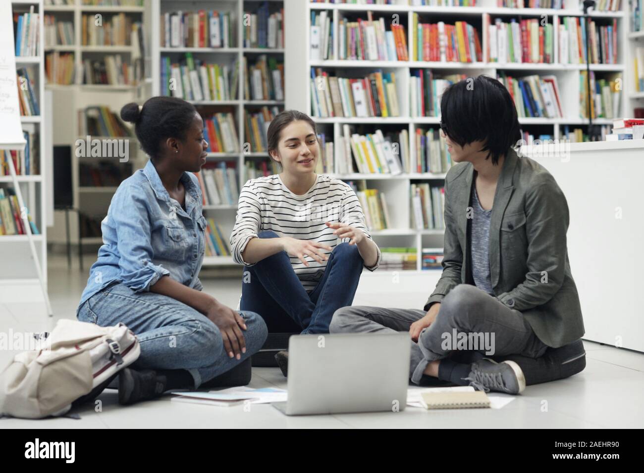 Multiethnic group of students sitting on the floor using laptop and discussing their homework in team in the library Stock Photo