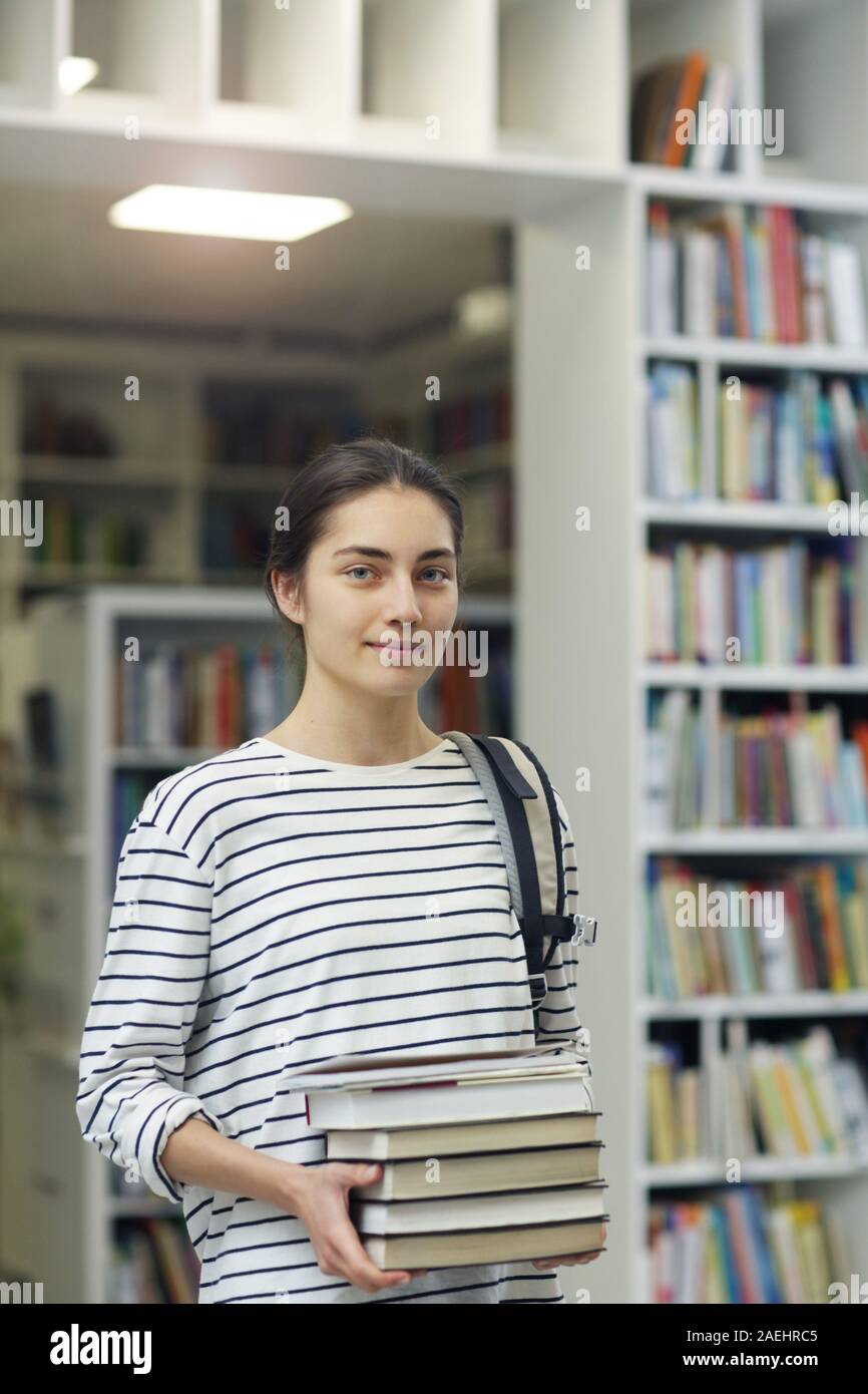 Portrait of young beautiful woman holding books and looking at camera while standing in the library Stock Photo