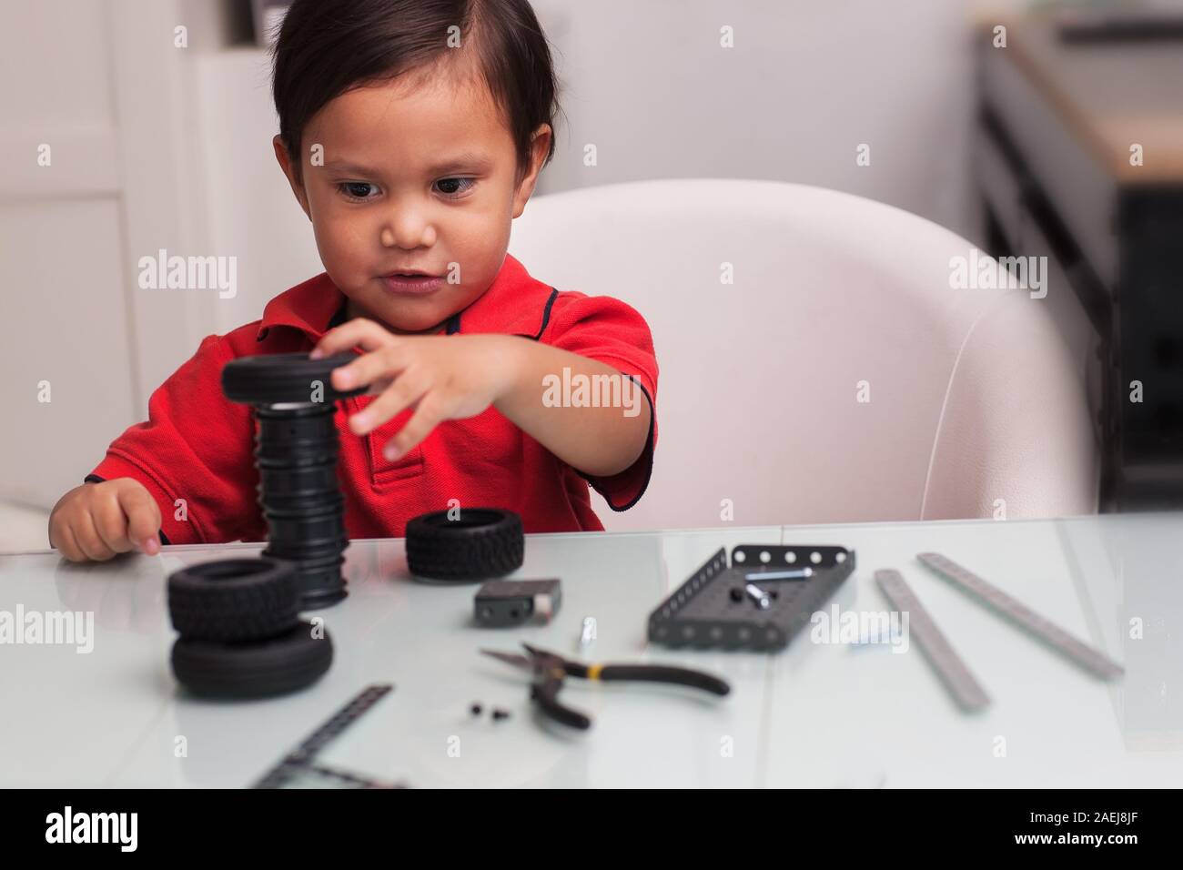 Three year old boy wearing a red shirt with fine motor skills is stacking toy tires on a glass desk with toy building parts. Stock Photo