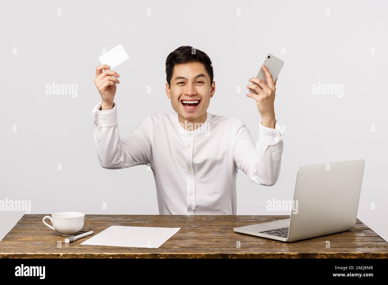 Man celebrating, hooray yes gesture, made good deal, ordered by best price. Asian businessman raising hands up, holding smartphone and credit card Stock Photo