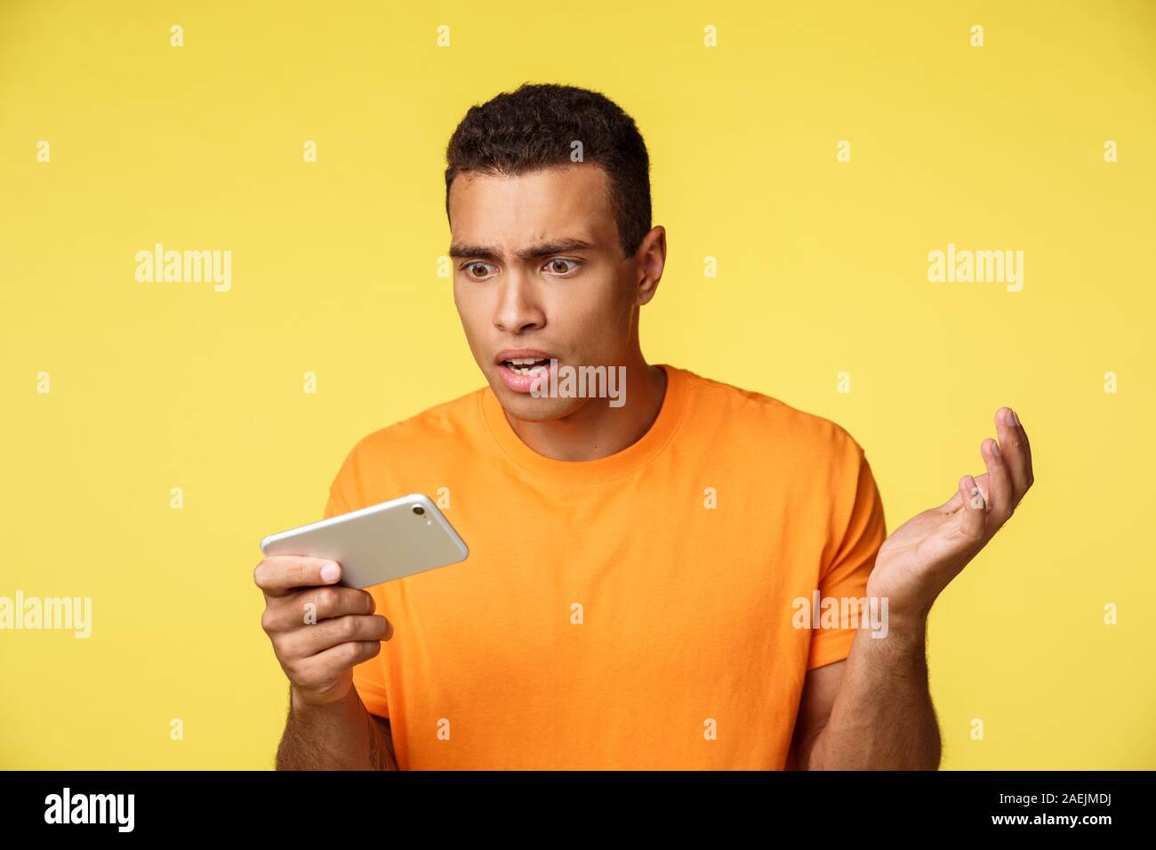 Shocked and bothered, upset handsome guy in orange t-sirt, stare at mobile screen questioned and sad, losing, see bad news, raise hands in dismay Stock Photo