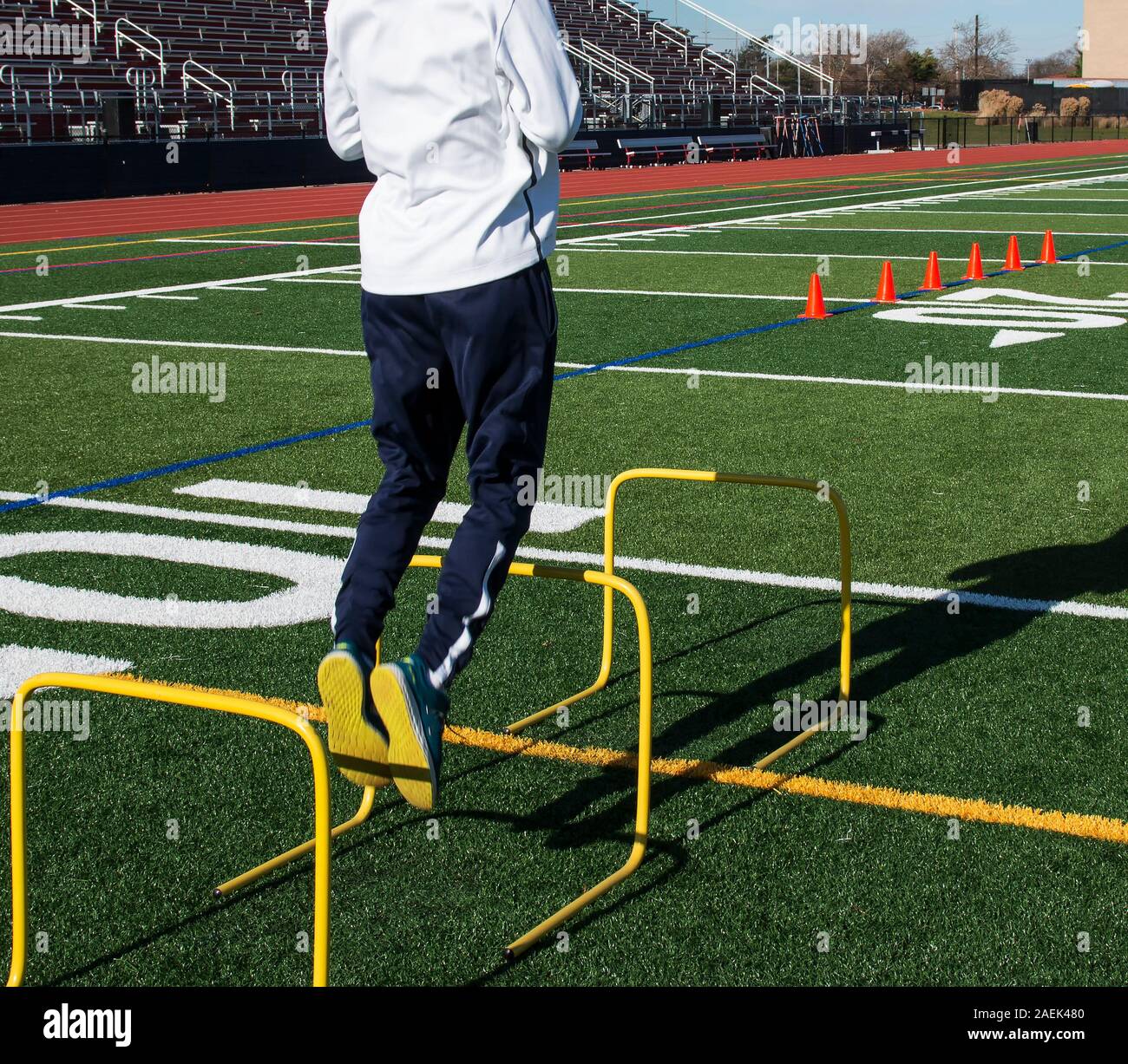 A track and field athlete jumping over yellow mini hurdles during cross training speed practice. Orange cones are in the background for running over. Stock Photo