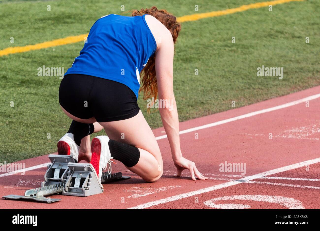 A high school female sprinter is in the starting blocks ready to start her sprint race on a red track outdoors. Stock Photo