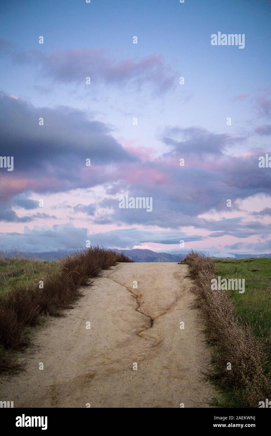 Dirt footpath on an undulating hill, disappearing over the horizon in the center of the image -taken at dusk with pink and blue sky and fluffy clouds. Stock Photo