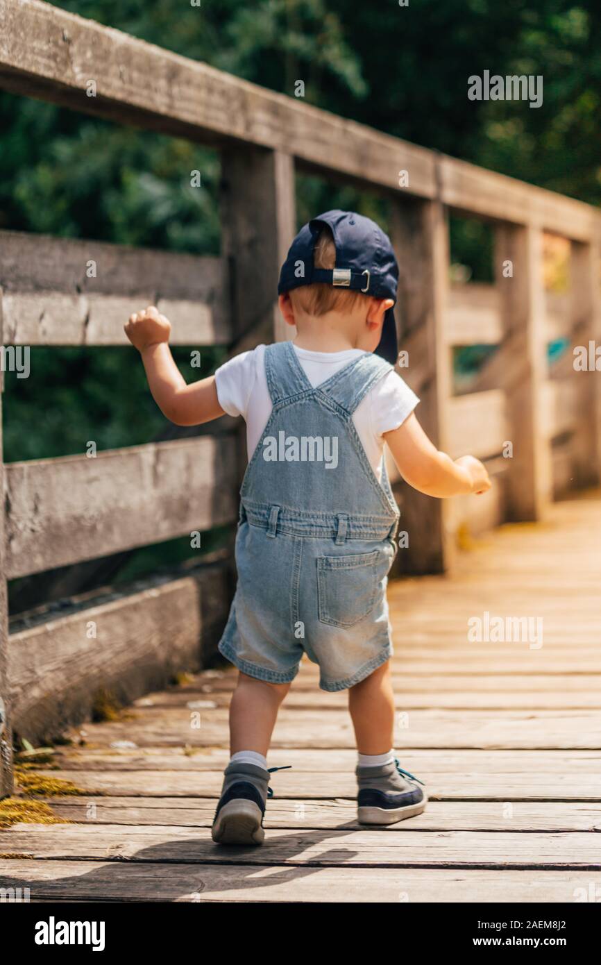 little child from behind walking on a wooden bridge in the alps with a blue baseball cap, hiking shoes and dungarees Stock Photo