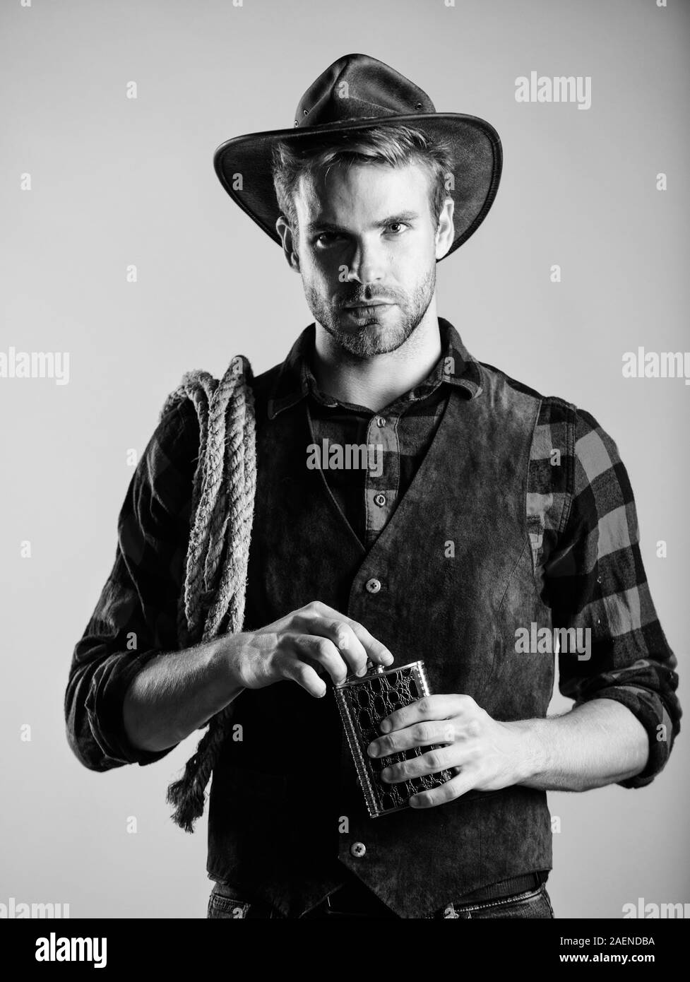 Bourbon whiskey. Western culture. Man wearing hat hold rope and flask. Lasso tool American cowboy. Sheriff concept. Brutal cowboy drinking alcohol. Man handsome cowboy beige background. Western life. Stock Photo