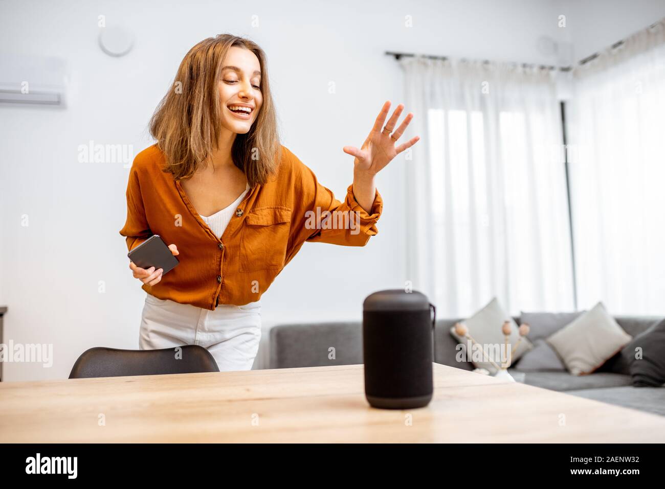 Young cheerful woman controlling home devices with a voice commands, talking to a smart column at home. Concept of smart home and voice command control Stock Photo