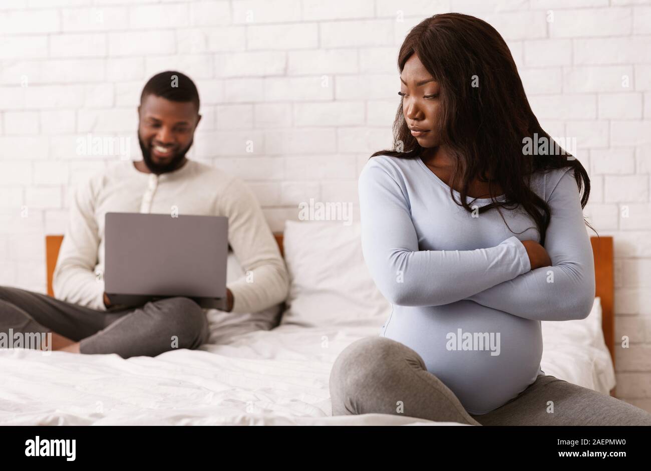 Stressed pregnant woman sitting on bed while husband using laptop Stock