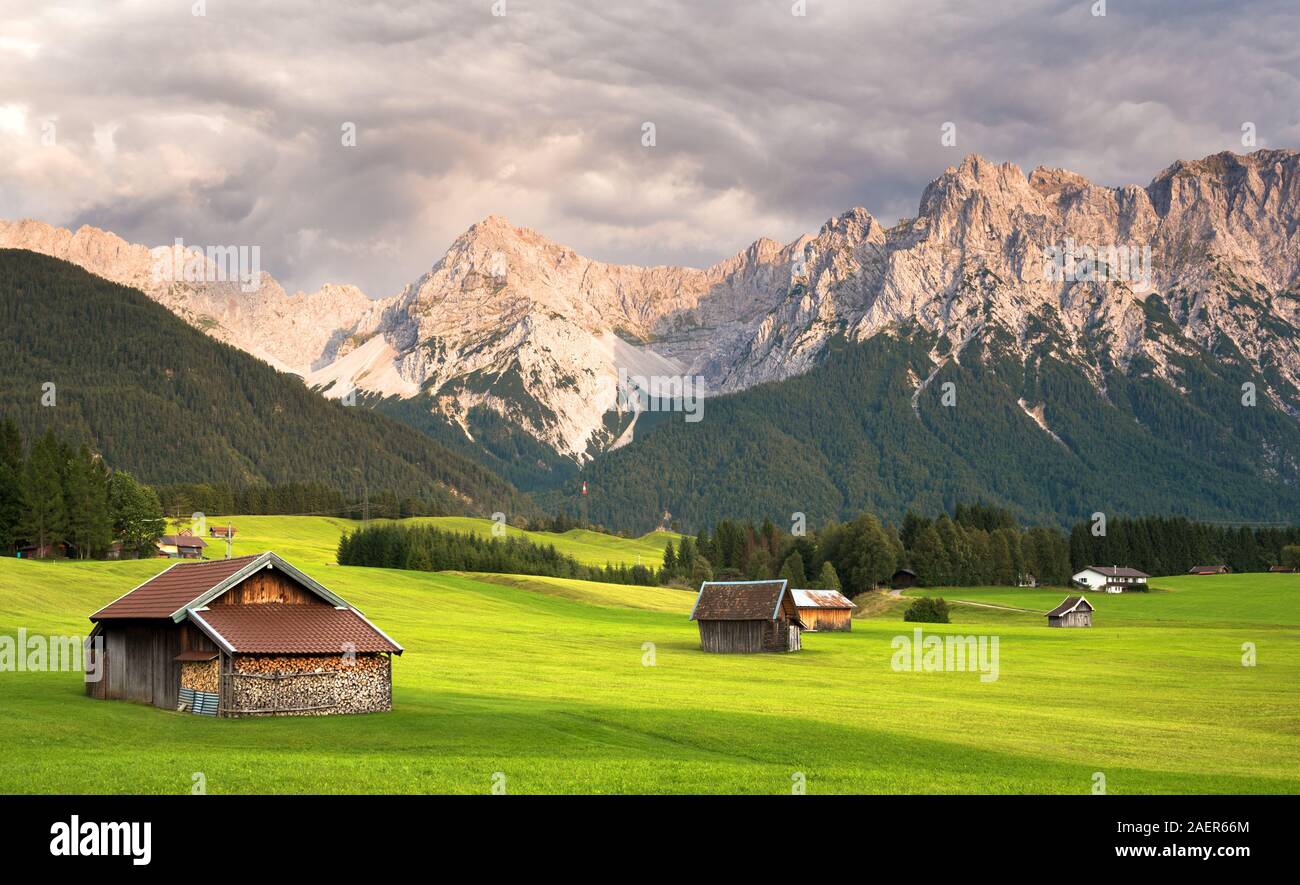 Wooden barns on sunlit green alpine meadow against mountain range and storm clouds Stock Photo