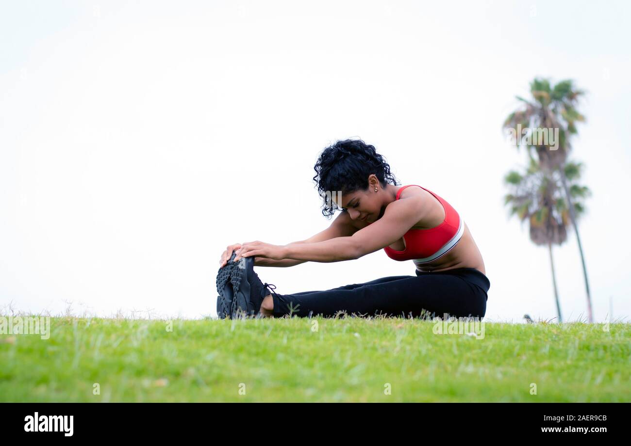 Fitness model performs hamstring stretching exercise by reaching and touching toes in city beach park Stock Photo