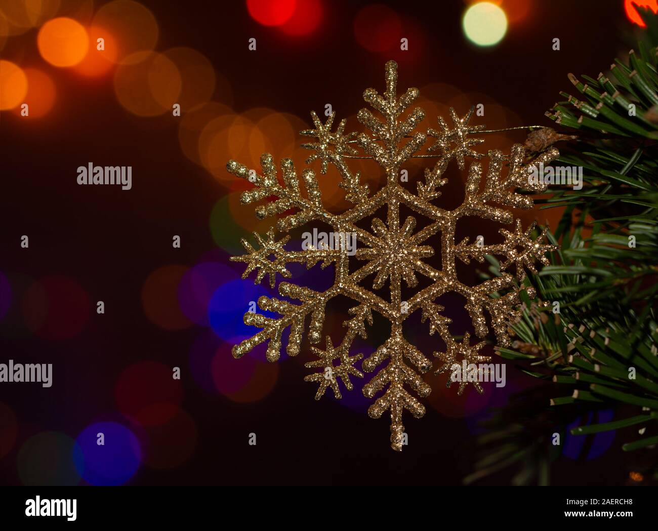 Gold colored glittery snowflake shaped Christmas ornament in tree, with bright colorful bokeh light background Stock Photo