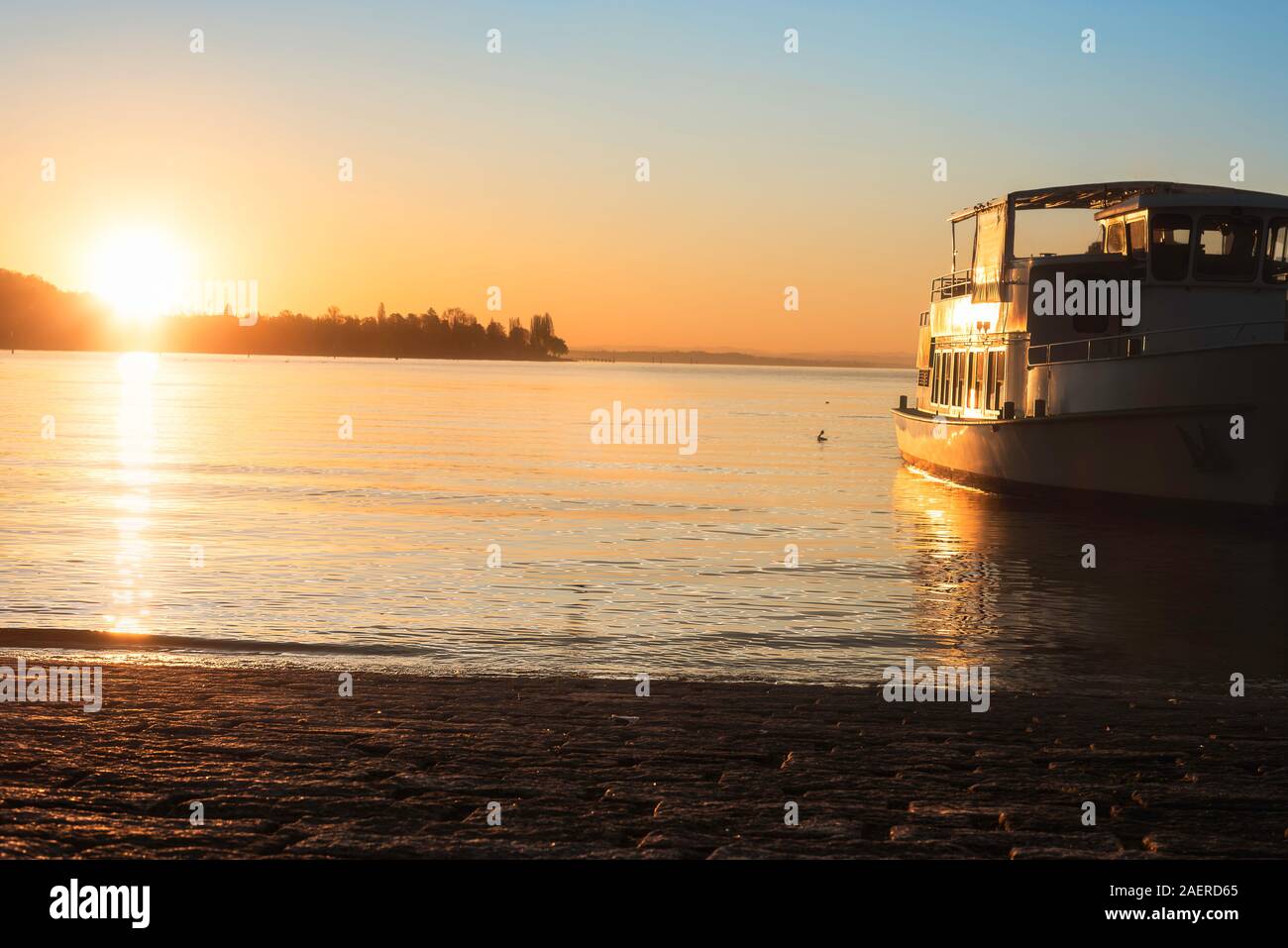 Sunrise over water and a boat on Constance lake, in Konstanz, Germany. Bodensee lakeshore and a ship cruising towards the sunrise. Water navigation. Stock Photo