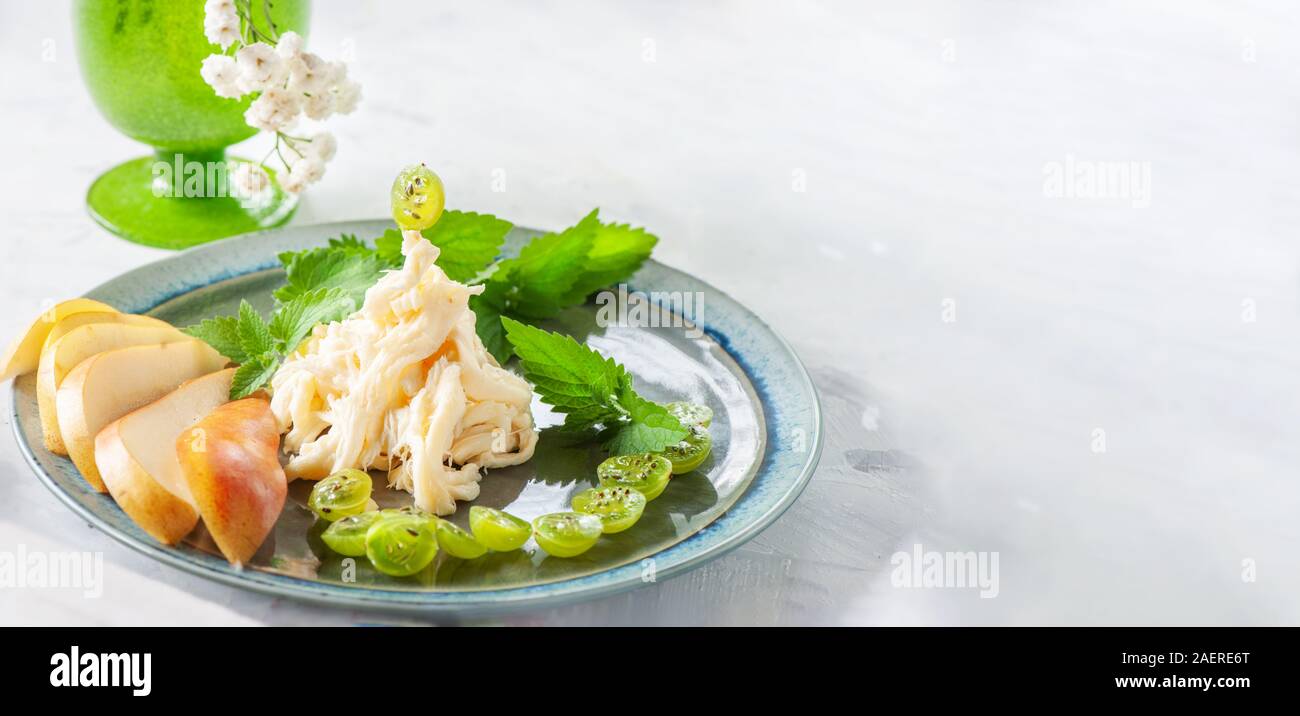 Traditional white soft fibrous Cecil cheese lies on a plate. The cheese is decorated with pieces of ripe pear and gooseberries. Copy space, banner. Stock Photo