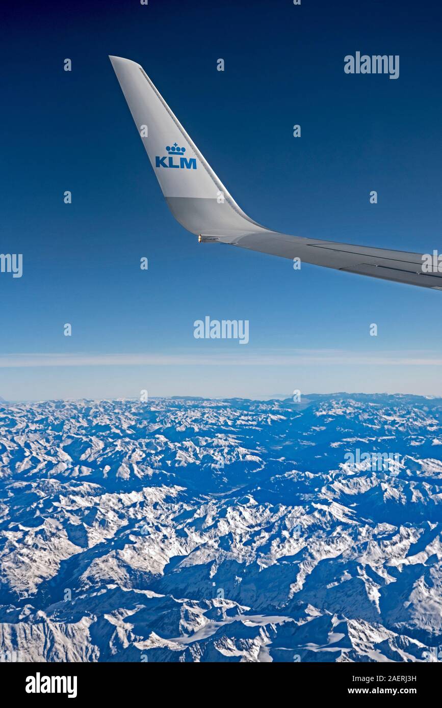 View from a KLM jet showing the wing with the KLM logo against a blue sky over the Alps. Stock Photo