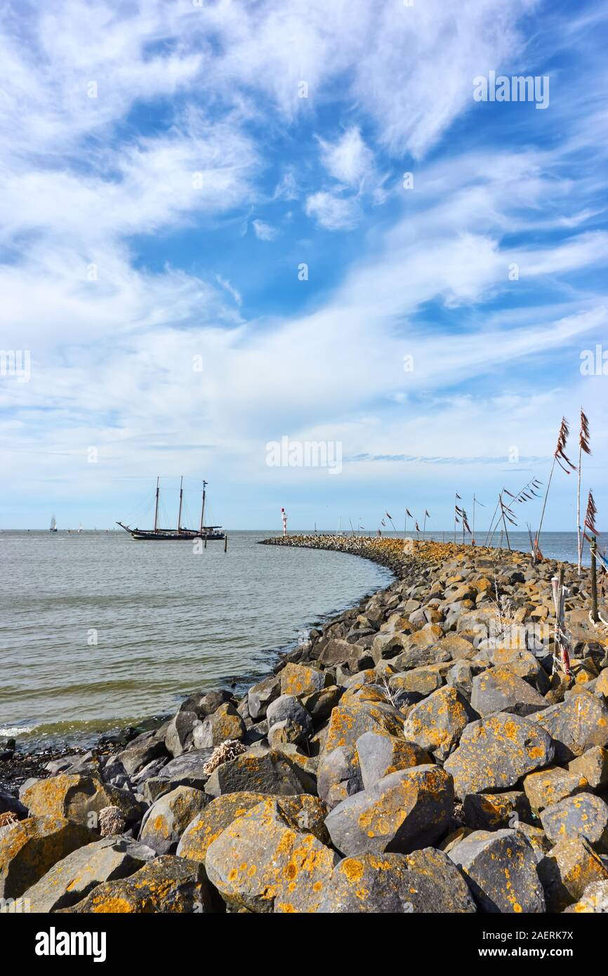 Vertical shot of breakwater or pier and poles with colored rope and plastic waving in the wind and a ship in the Wadden Sea. Stock Photo