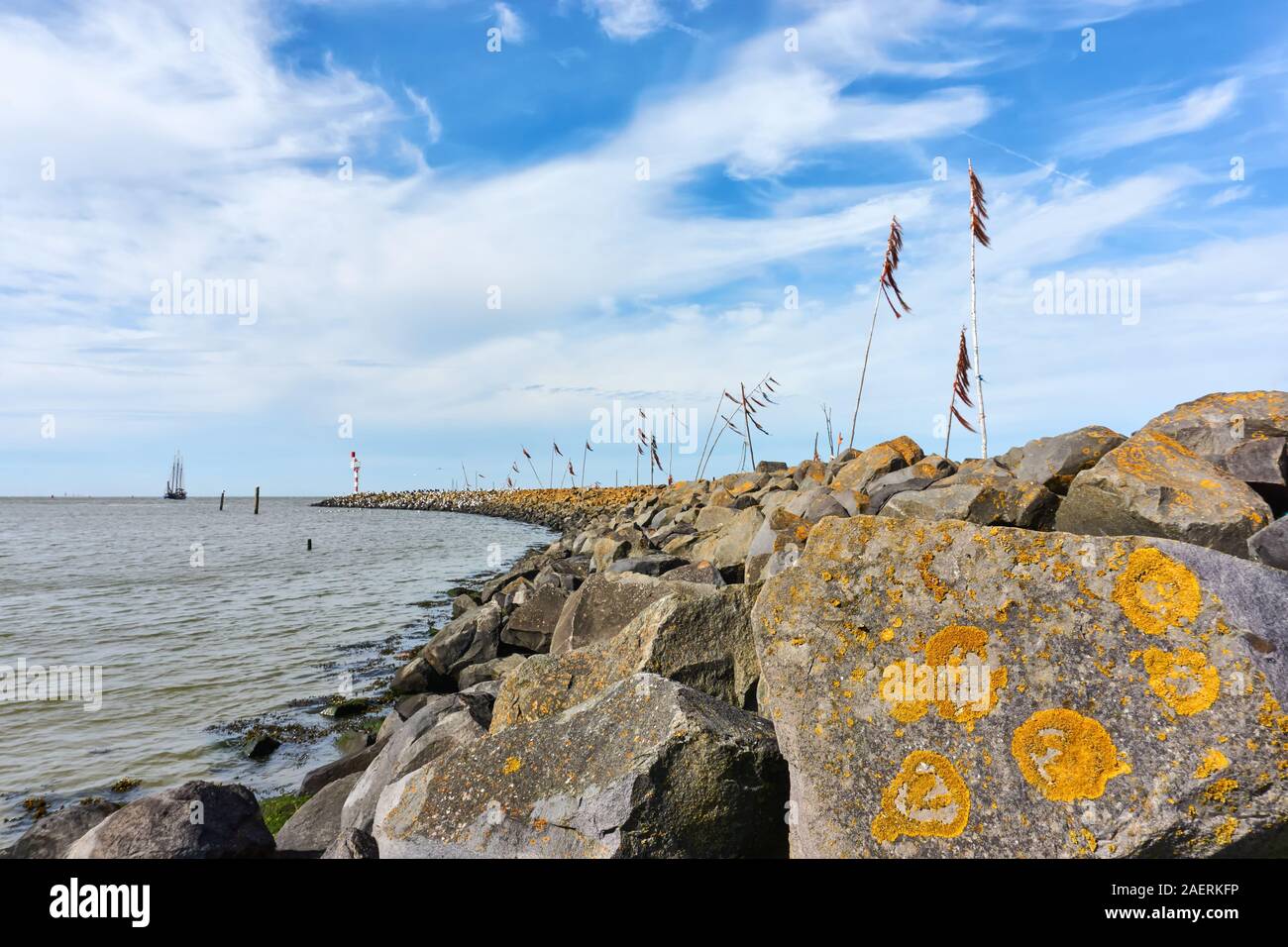 Breakwater or pier and poles with colored rope and plastic waving in the wind and a ship in the Wadden Sea at the entrance to the port of Harlingen Stock Photo