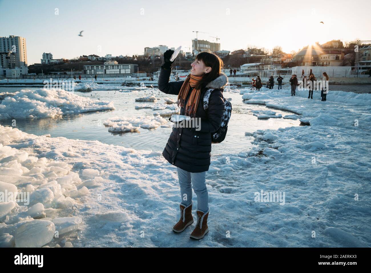 Odessa, Ukraine - February, 16 2017: Young smiling woman standing on frozen coast of the Black sea covered with ice holding piece of ice at sunset in Stock Photo