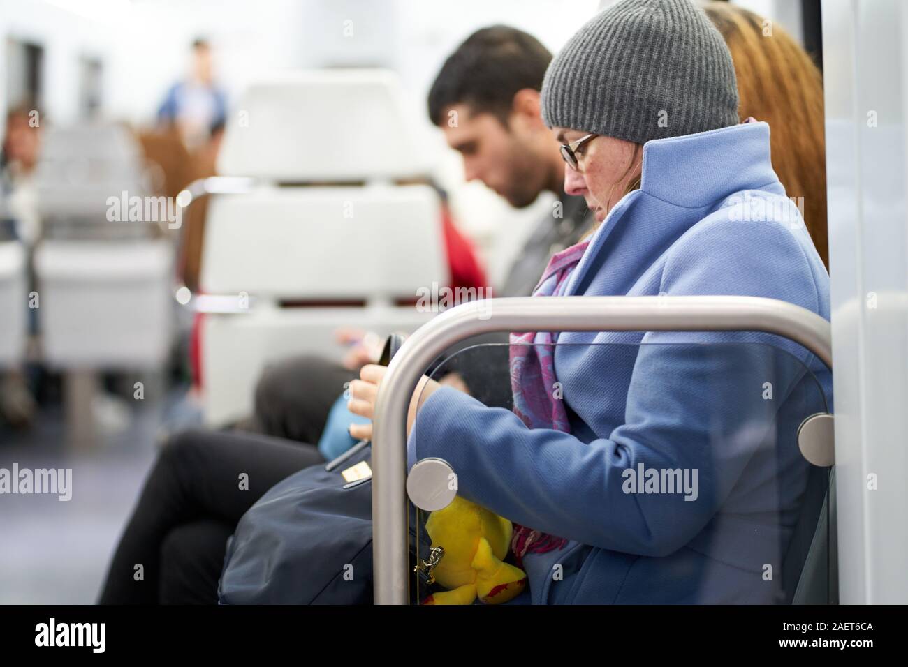 A young girl, sitting on a train in a blue coat with a beanie hat and glasses attends to her bag whilst holding a yellow duck Stock Photo