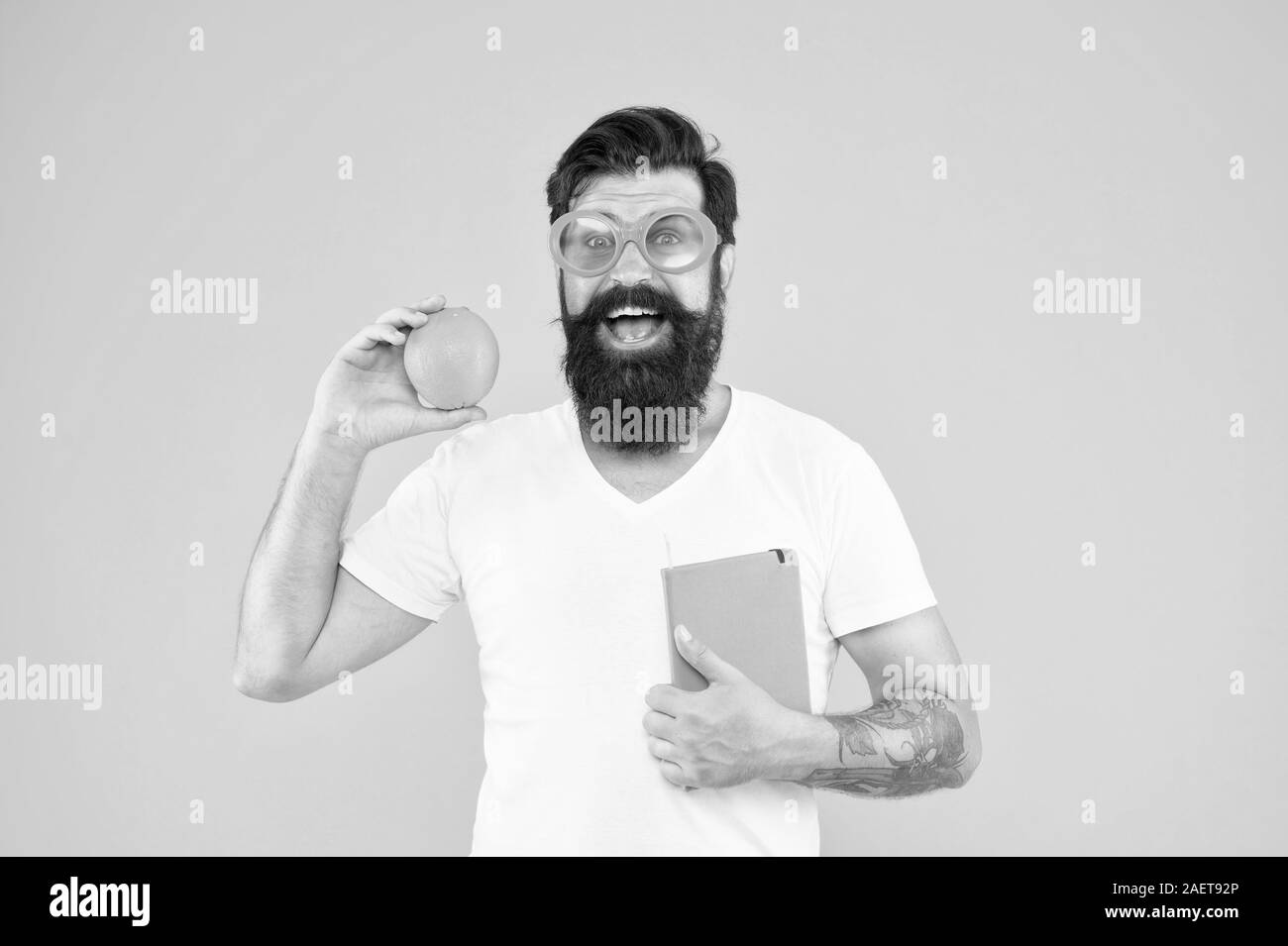 Nerd is the new cool. Bearded nerd man. Study nerd holding book and orange fruit on yellow background. Book nerd in fancy glasses choosing natural diet. Stock Photo