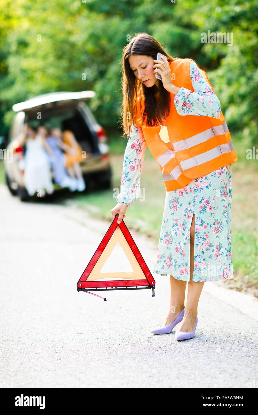 Woman in orange reflective vest installing red warning triangle on the road, children and broken car in background Stock Photo