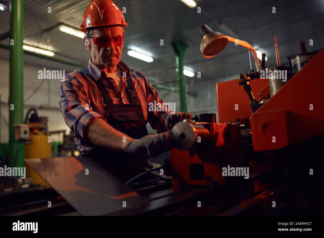 Serious mature manual worker in protective work wear standing at the lathe and making metal details Stock Photo