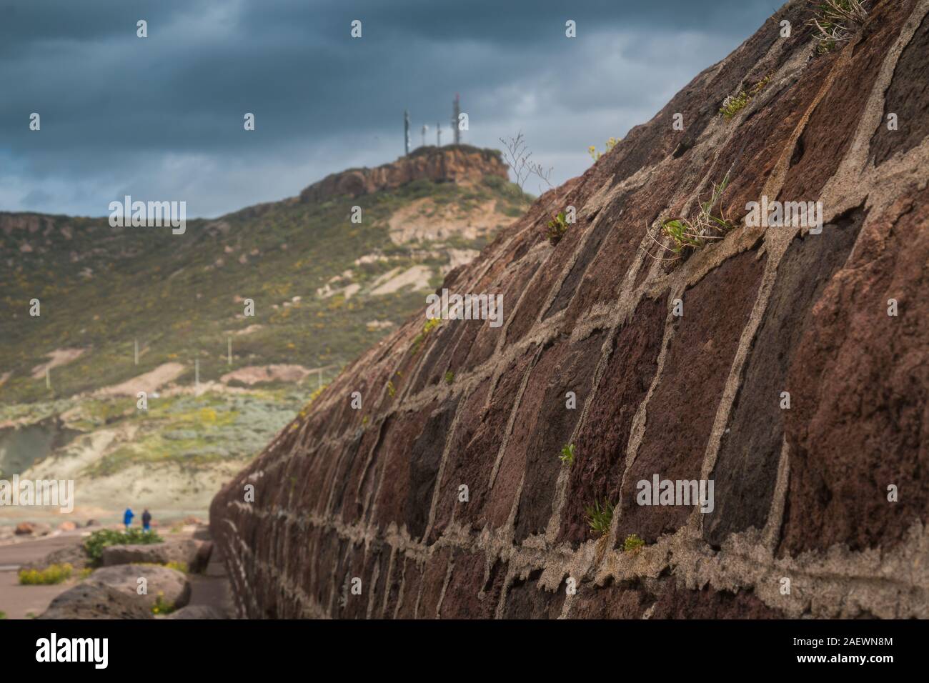 Detail of a high wall made of stones, between the sea and an old tower. Mountain in the background. Intense stormy clouds. Bosa Marina, Sardinia, Ital Stock Photo