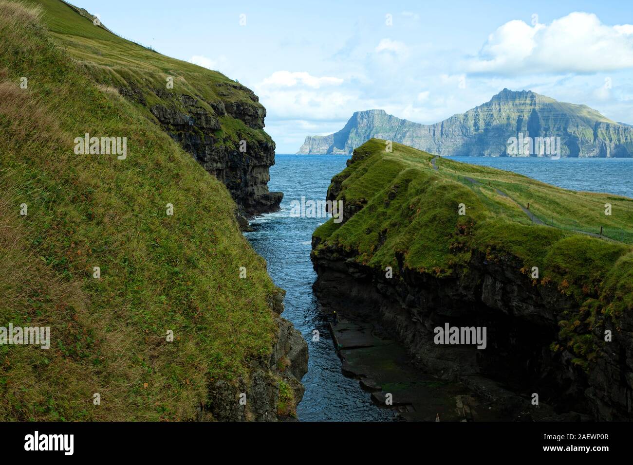 Direct view into the gorge / natural harbour of Gjogv with dramatic cloudy sky, lush green grass and turquoise water Stock Photo