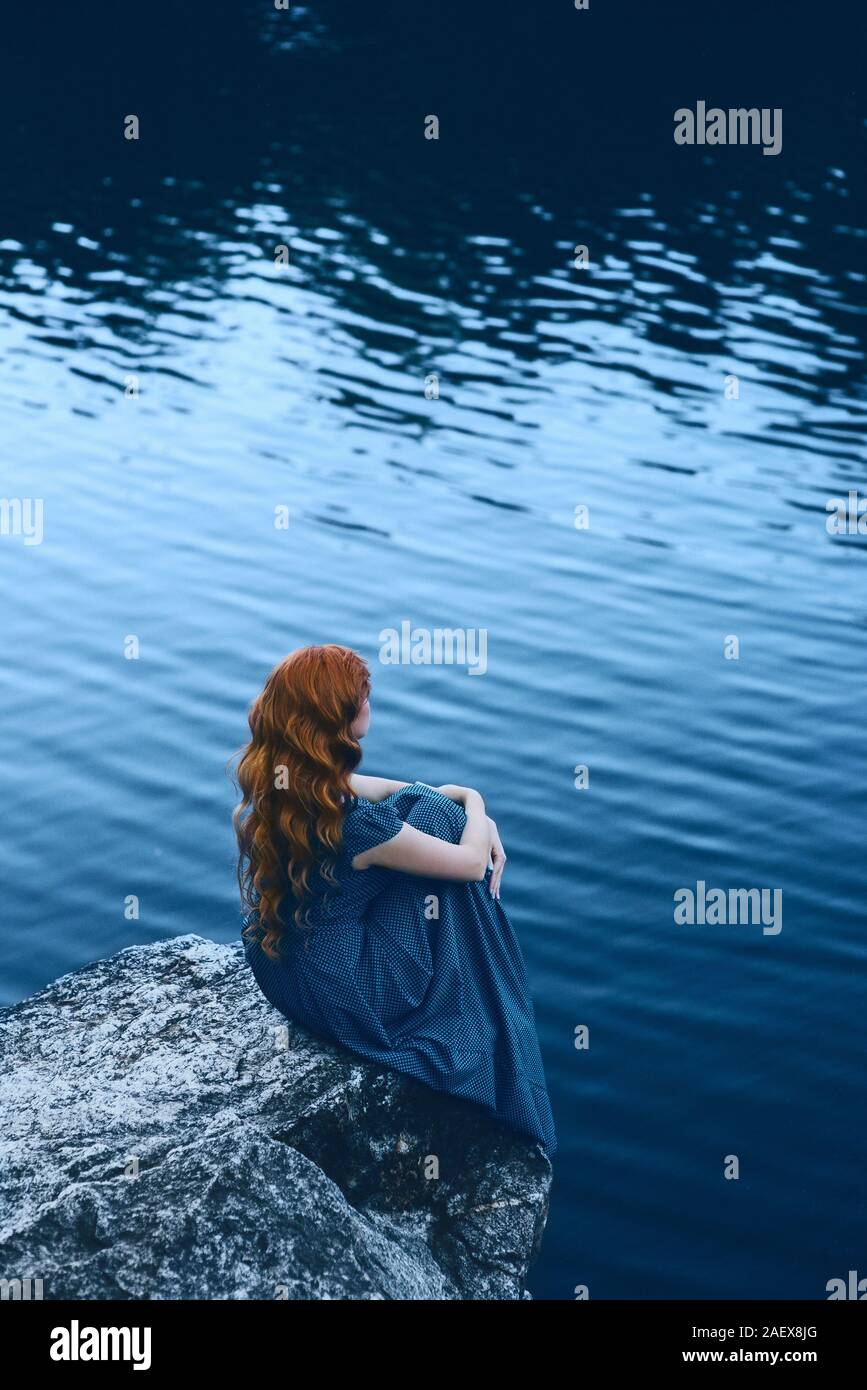 Beautiful woman in blue dress with long red hair sitting on a cliff by ...