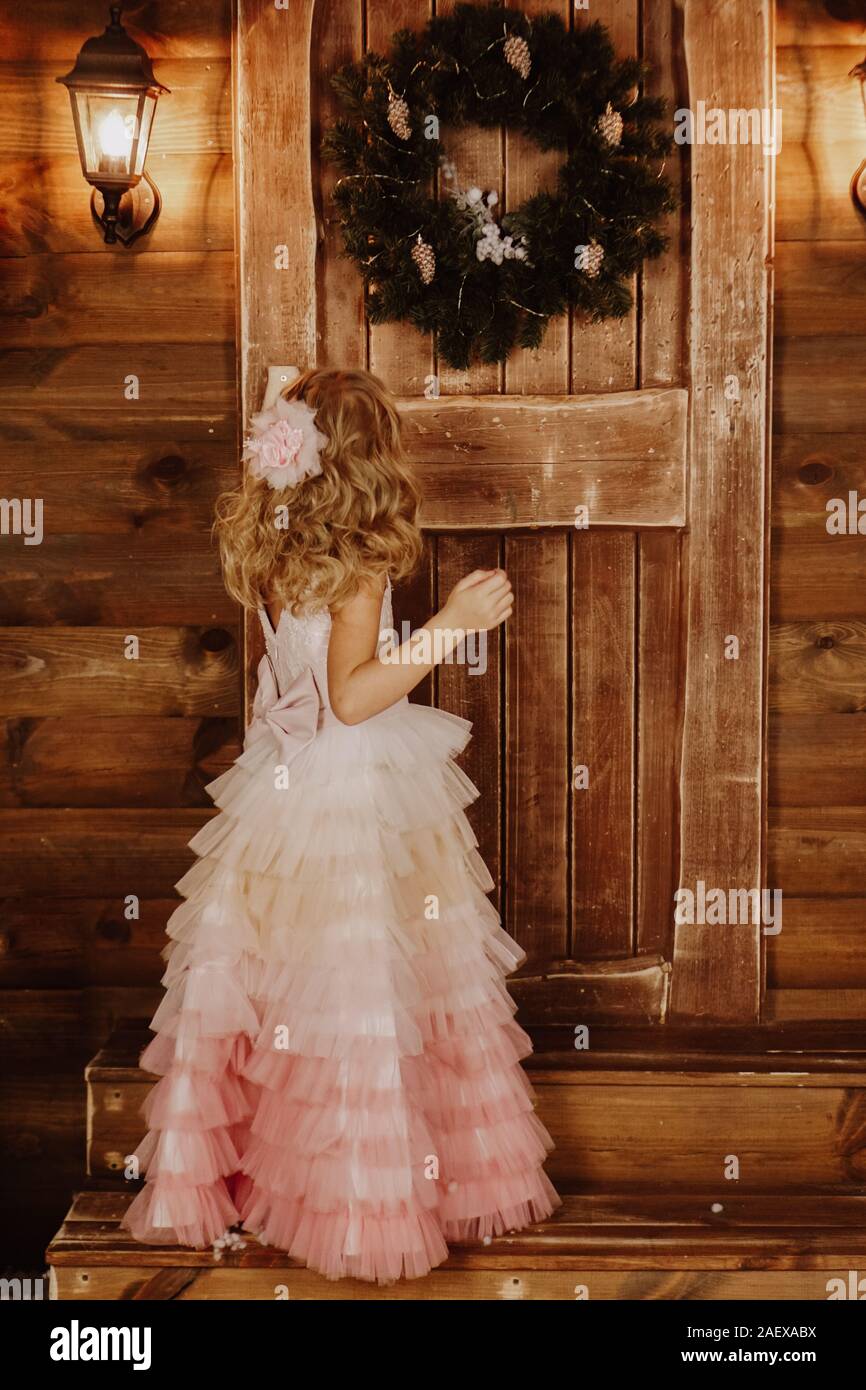 A little girl in a pink dress stands in front of a wooden door with a ...
