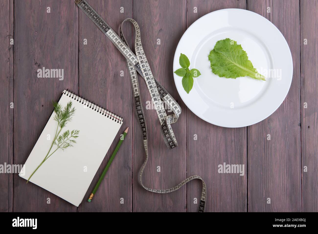 Healthy diet plan concept - blank notepad for recipe, measure tape, white plate with dill, salad lettuce, basil on wooden table Stock Photo