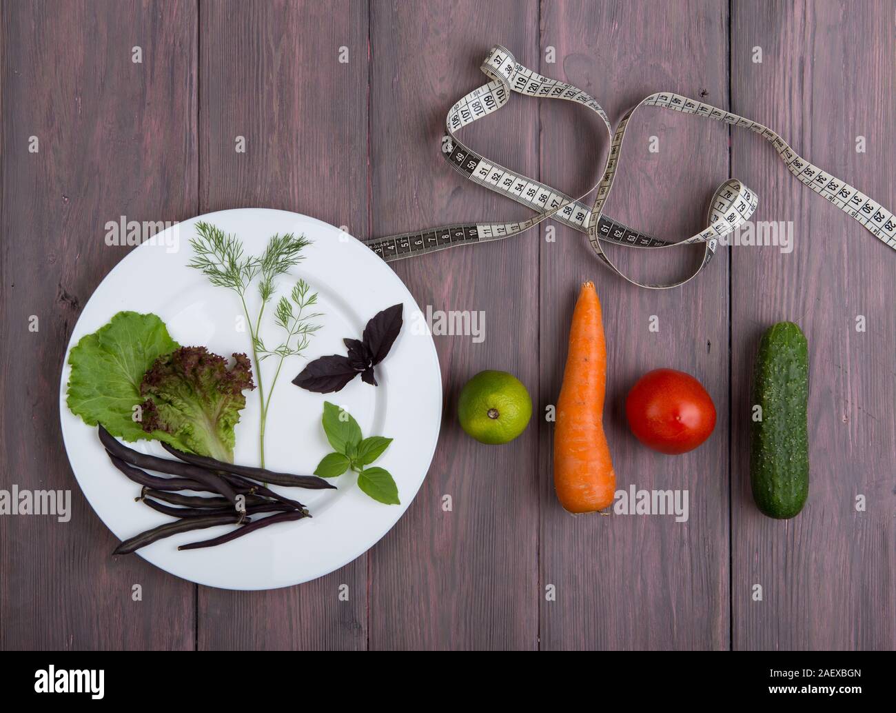 Healthy food and diet vegetarian concept - measure tape, white plate with leaf of salad, cucumber, tomato and other vegetables on wooden table Stock Photo