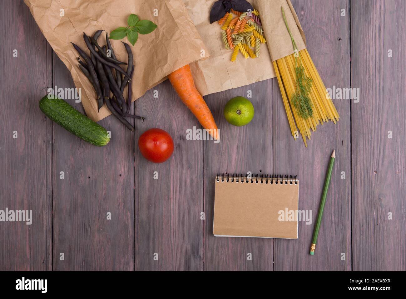 Fresh pasta ingredients in eco paper bags - spaghetti pasta, carrot, basil, tomato and other vegetables, notepad for recipe on wooden table Stock Photo