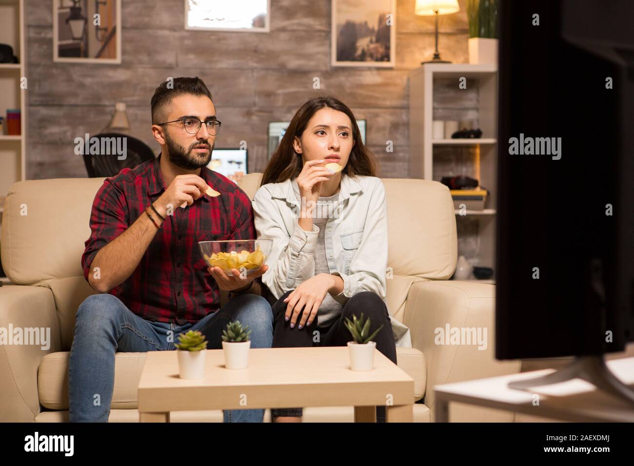 Beautiful young couple looking worried at tv while watching a movie. Couple eating chips sitting on sofa. Stock Photo