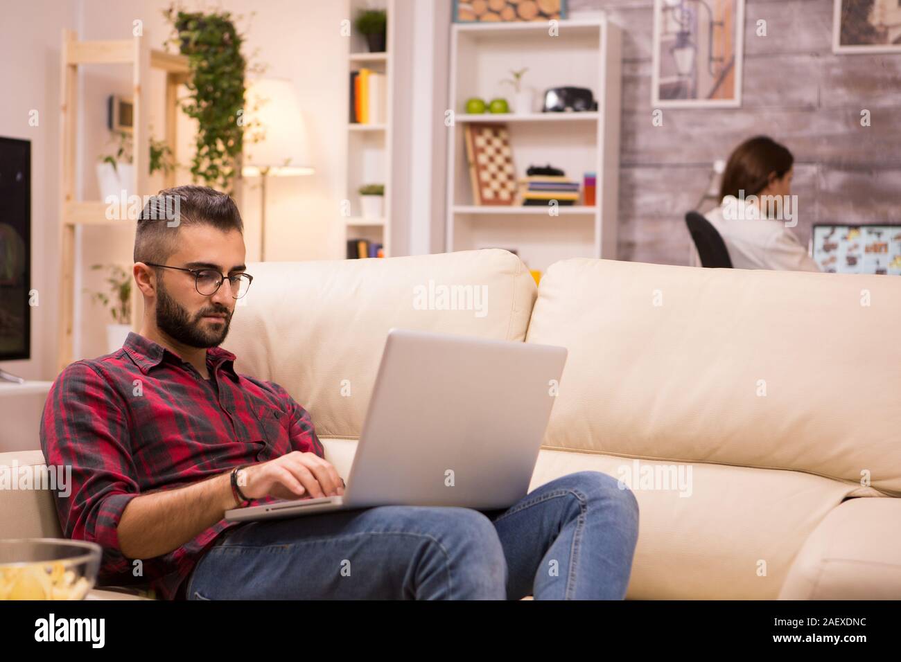 Young entrepreneur working on laptop sitting on sofa. Girlfriend in the background. Stock Photo