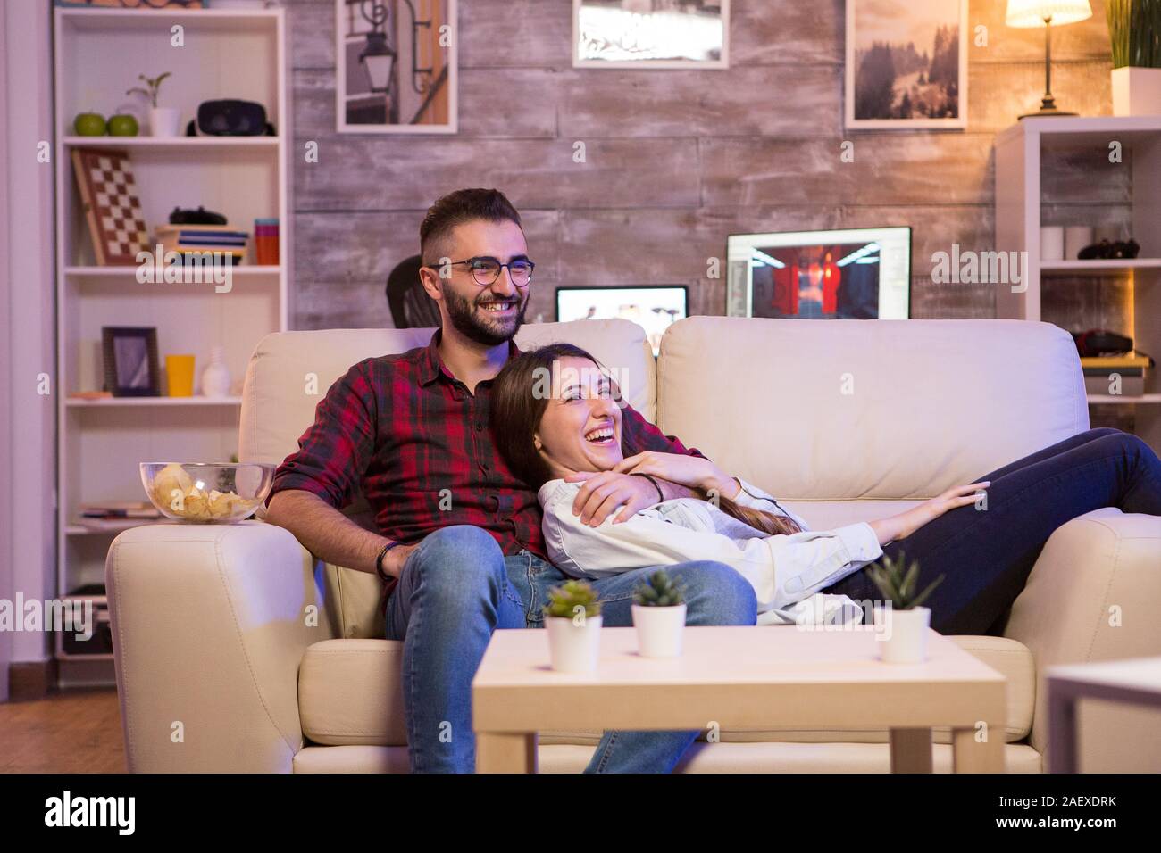 Cheerful young couple laughing while watching a tv show on tv at night. Couple sitting on sofa. Stock Photo