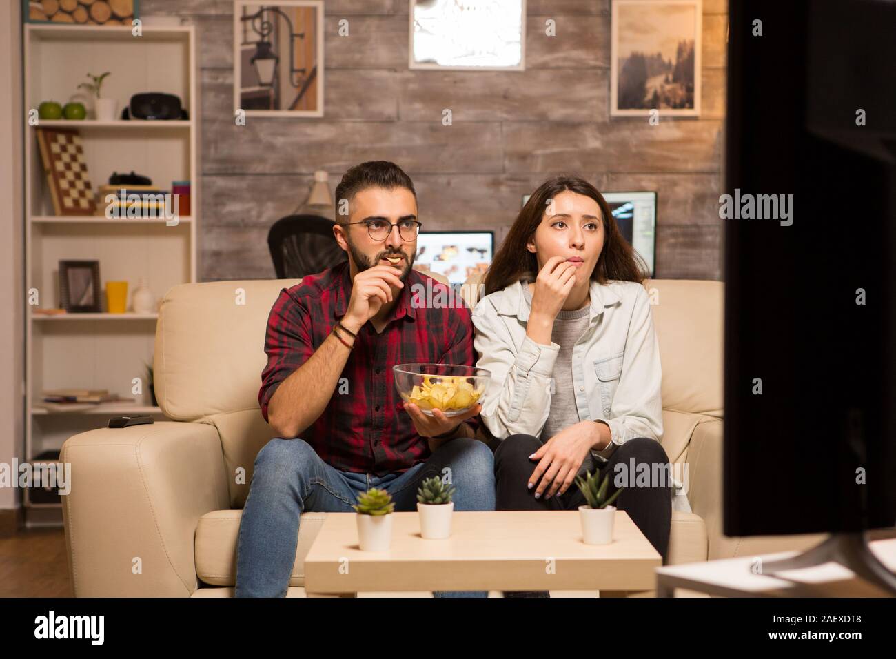 Young couple looking concentrated on tv while watching a movie and eating chips . Couple sitting on sofa. Stock Photo