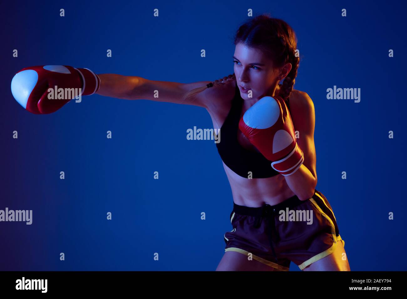 Fight. Fit caucasian woman in sportswear boxing on blue studio background in neon light. Novice female caucasian boxer working out and training. Sport, healthy lifestyle, movement concept. Stock Photo