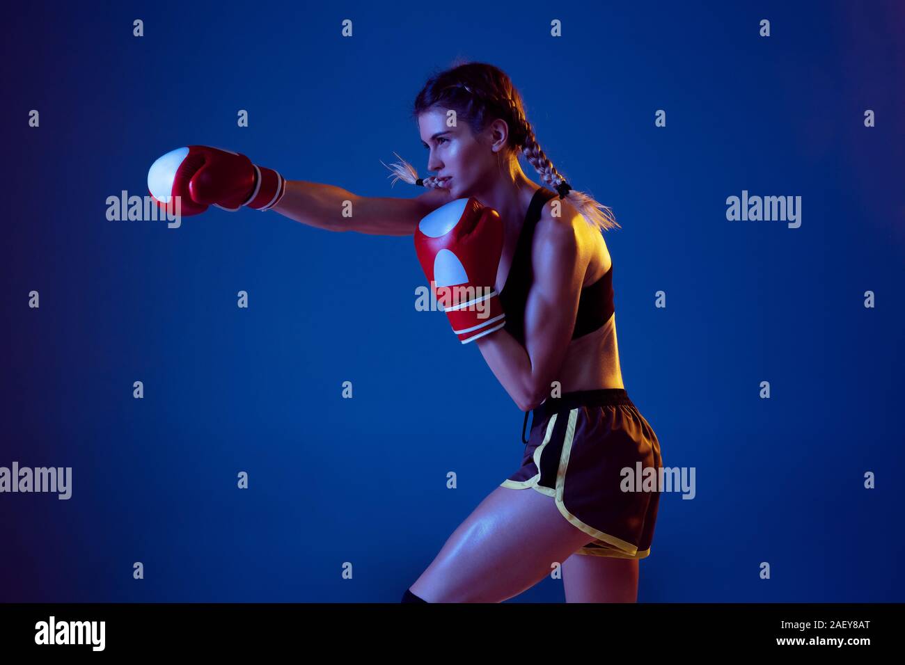 Beautiful. Fit caucasian woman in sportswear boxing on blue studio background in neon light. Novice female caucasian boxer working out and training. Sport, healthy lifestyle, movement concept. Stock Photo