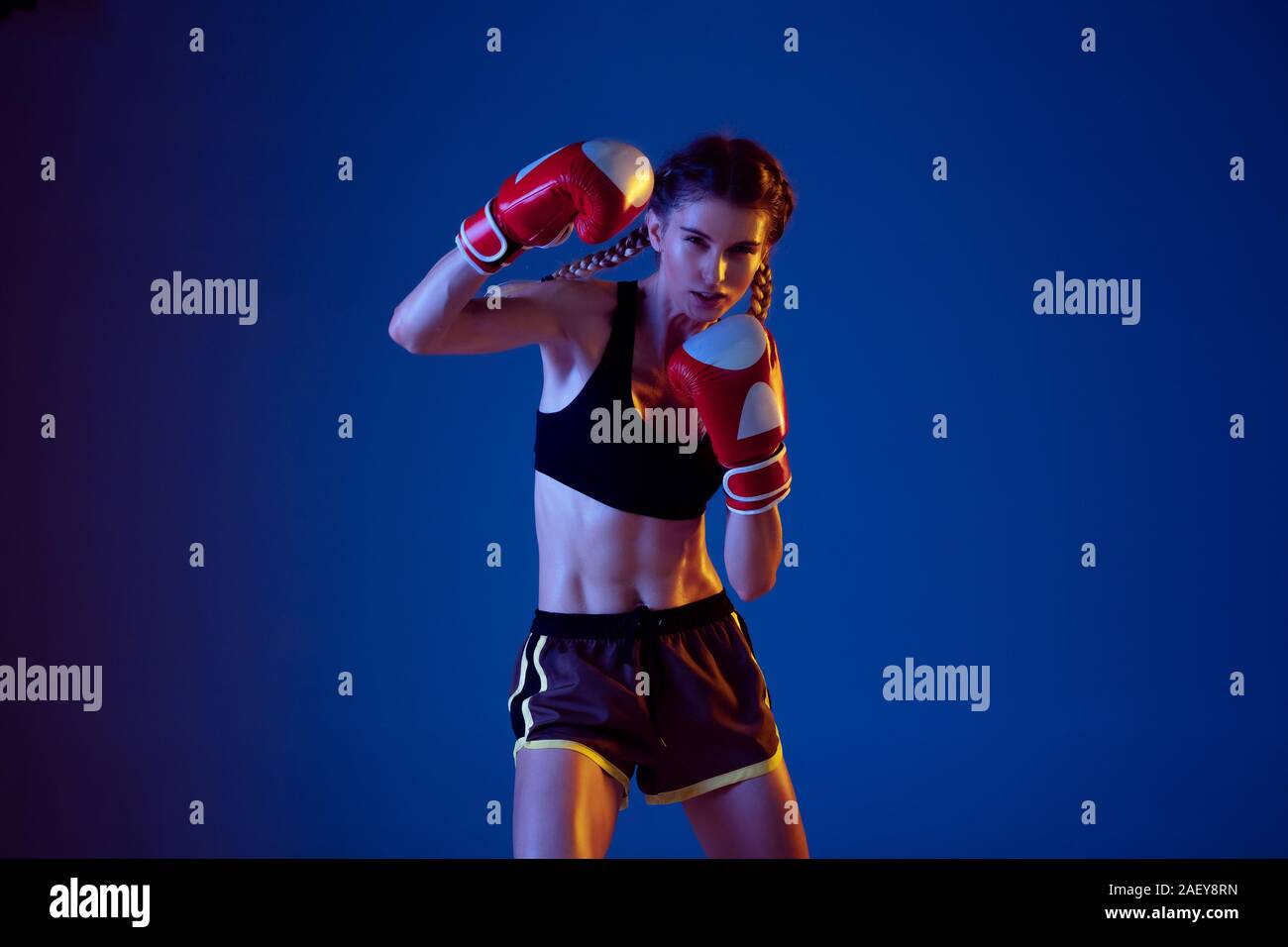 Fit caucasian woman in sportswear boxing on blue studio background in neon light. Novice female caucasian boxer working out and training. Sport, healthy lifestyle, movement concept. Copyspace. Stock Photo