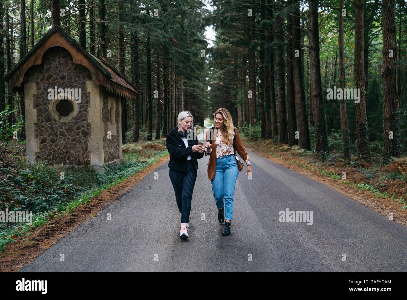 Two women walking on a road in the forest while watching their phone Stock Photo