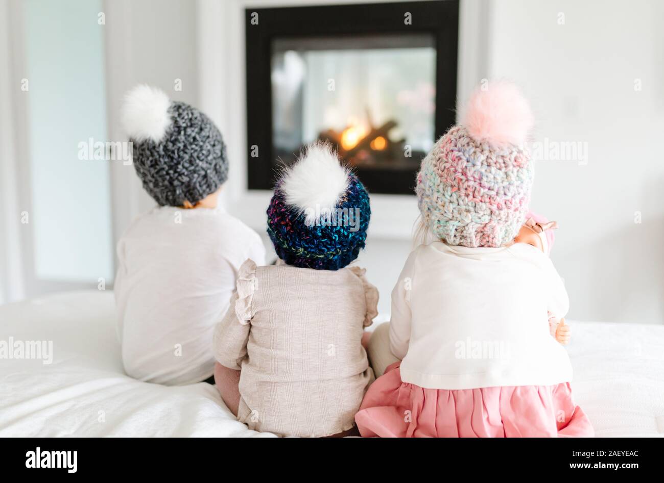 Three toddlers sitting on bed in front of fireplace with knit hats Stock Photo