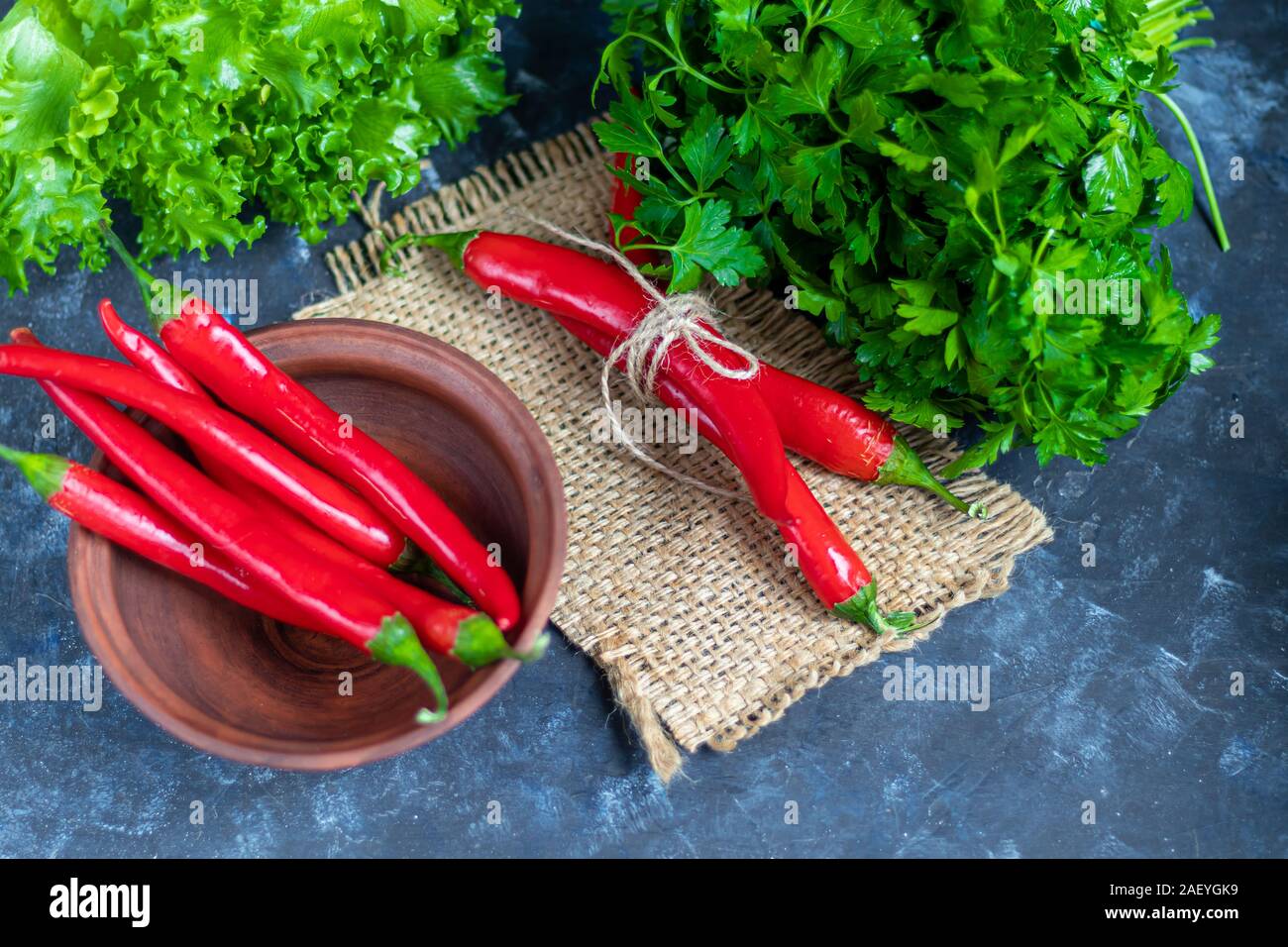 Hot red chili peppers on a dark background under concrete. Spicy food supplement. Parsley and lettuce leaves. Copy space. Stock Photo
