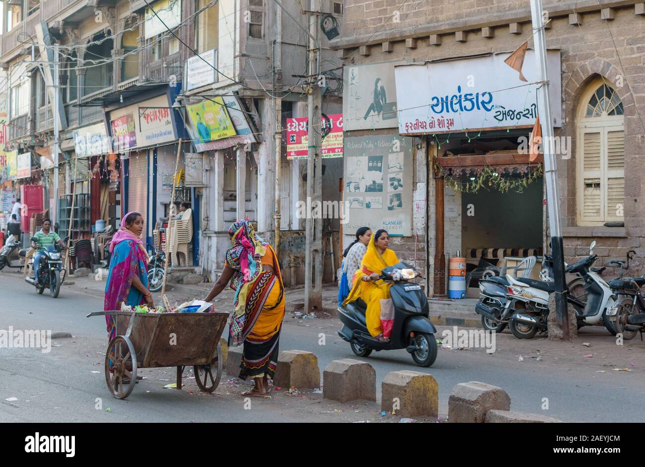 Two woman pick garbage off the streets in the middle of the traffic in the old town market. Gujarati text on signboards outside shops. Stock Photo