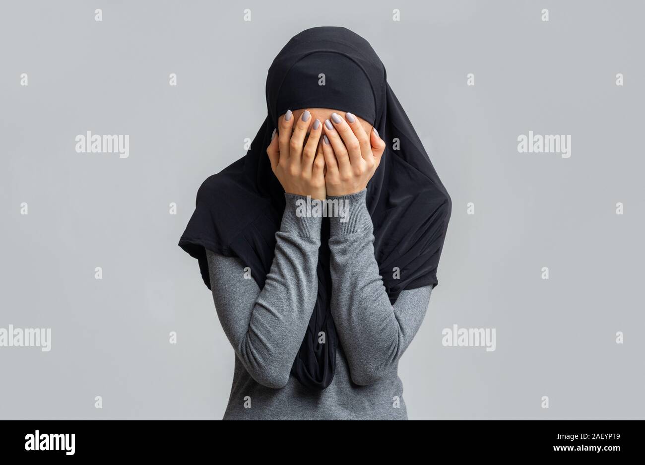Islamophobia and muslim women discrimination concept. Woman in black hijab covering face with hands, posing over gray studio background Stock Photo