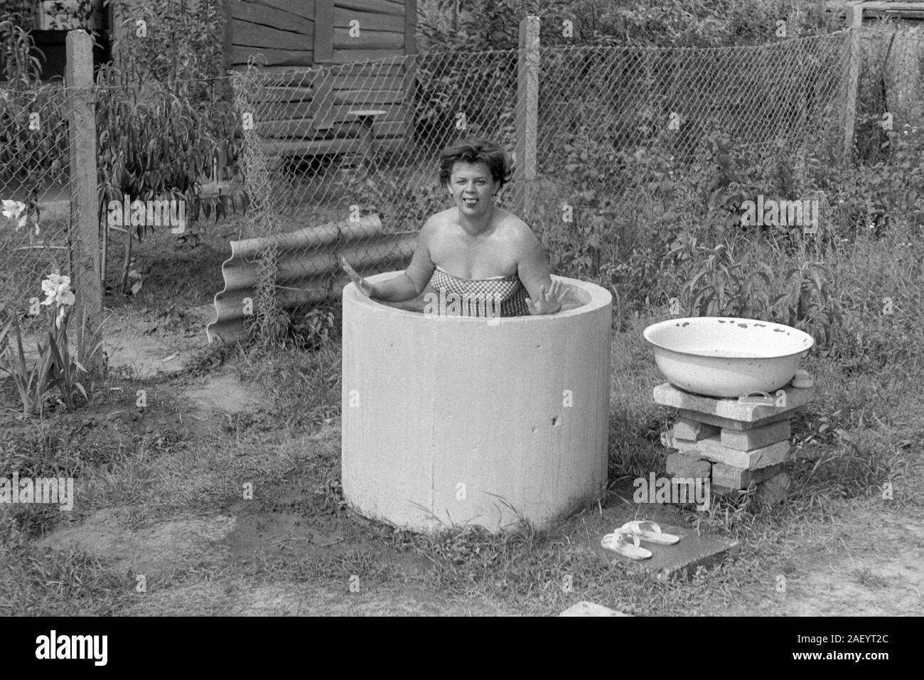 woman in bathing costume bathes in garden using improvised pool made from a section of drain pipe using water heated on an open fire 1960s hungary Stock Photo